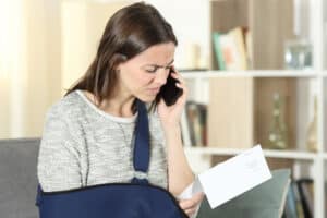 woman-with-arm-injury-sling-on-phone-reading-paperwork-confused